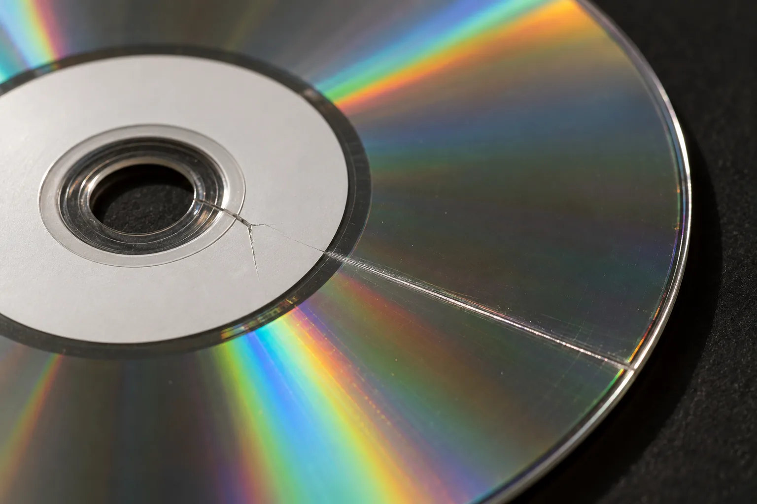 Close-up van een cd op een donkere ondergrond met een haarscheurtje in het plastic aan de rand van het middengat en een rechte kras die van daar naar de buitenrand loopt.